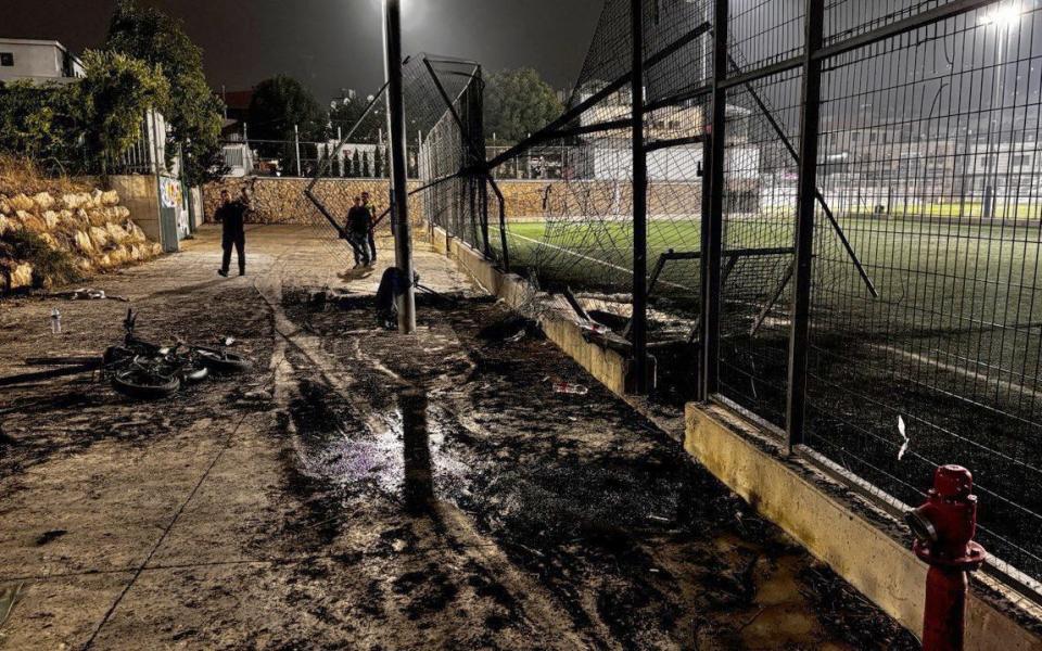 The area next to the pitch is blackened with ashes, a chain-link fence is twisted and destroyed and a child's bicycle lies abandoned