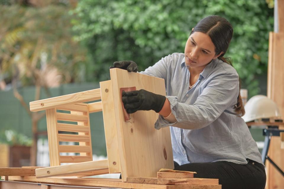 Female carpenter making a little wooden table in her workshop. Young female using sandpaper doing woodwork in a warehouse for her small furniture or carpentry business from her home.