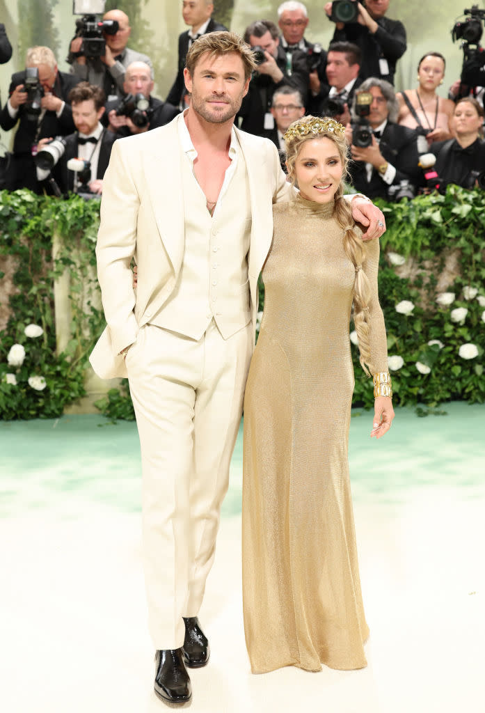 Chris Hemsworth in a  suit and Elsa in a gold dress pose together on a greenery-backdropped event