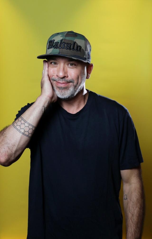 A man in a black T-shirt and camouflage baseball cap stands, his hand along the side of his face.