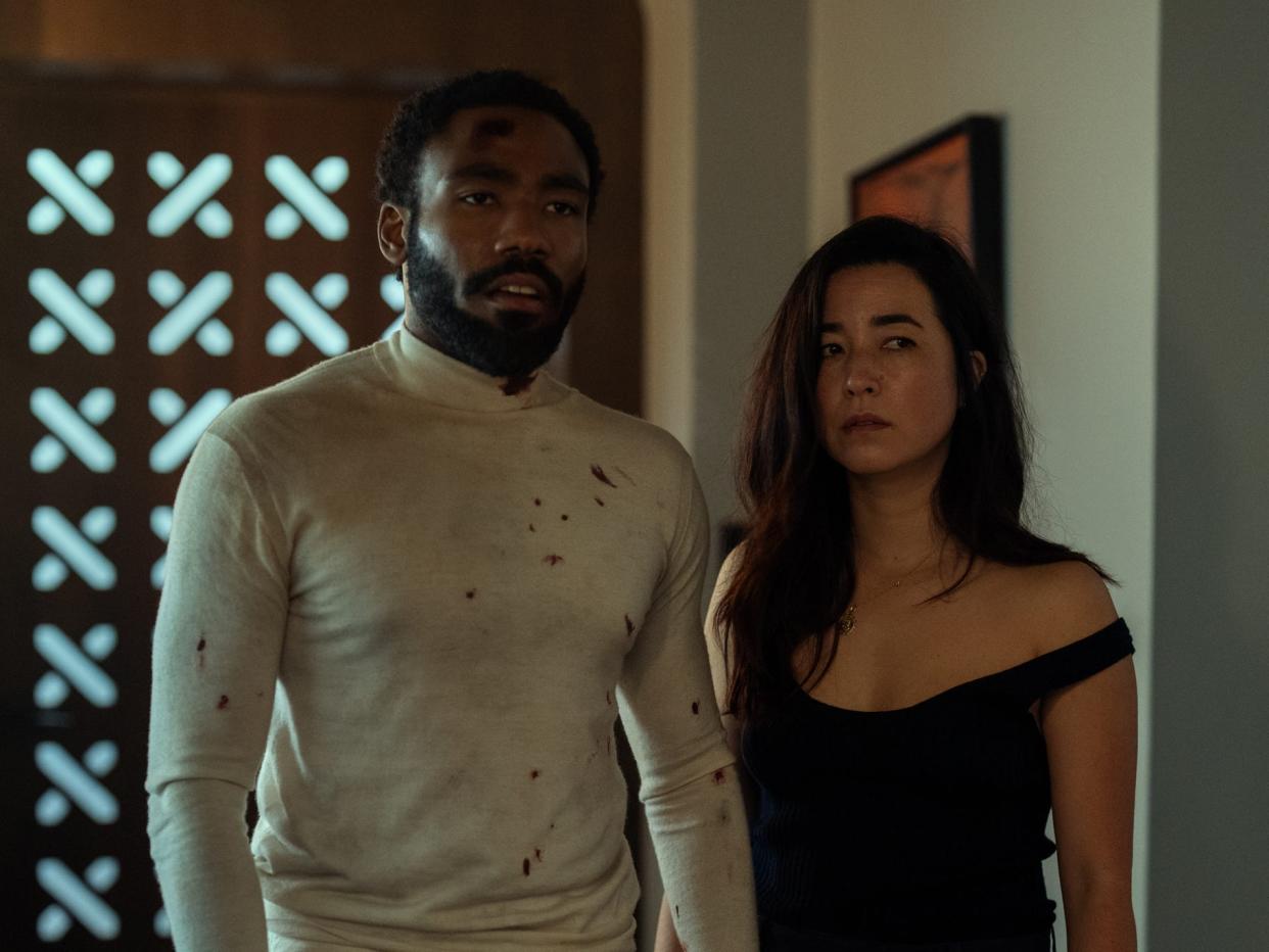 donald glover and maya erskine in mr. and mrs. smith. glover is wearing an all white outfit splatterd with blood and grime. erskine is in a black outfit that's in disarray