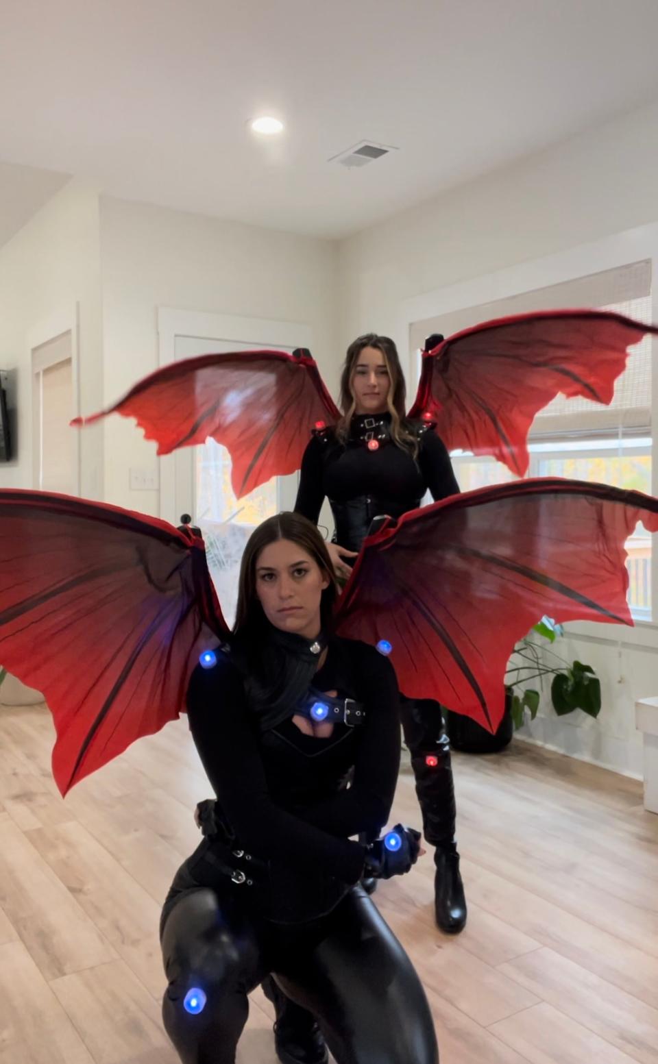 Two women dressed in black suits with red wings pose.