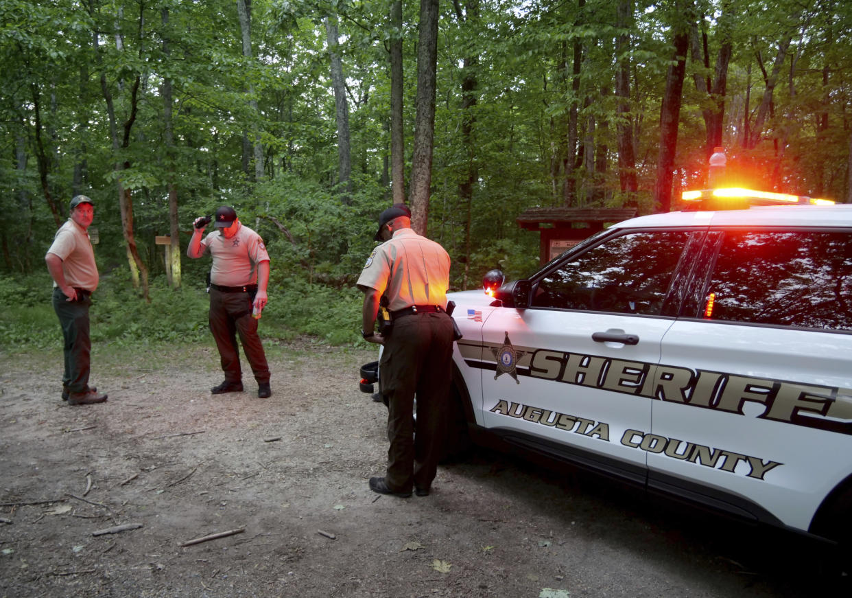 Three service members stand in what appears to be a clearing in the woods, near a patrol car with lights flashing emblazoned with: Sheriff, Augusta County  