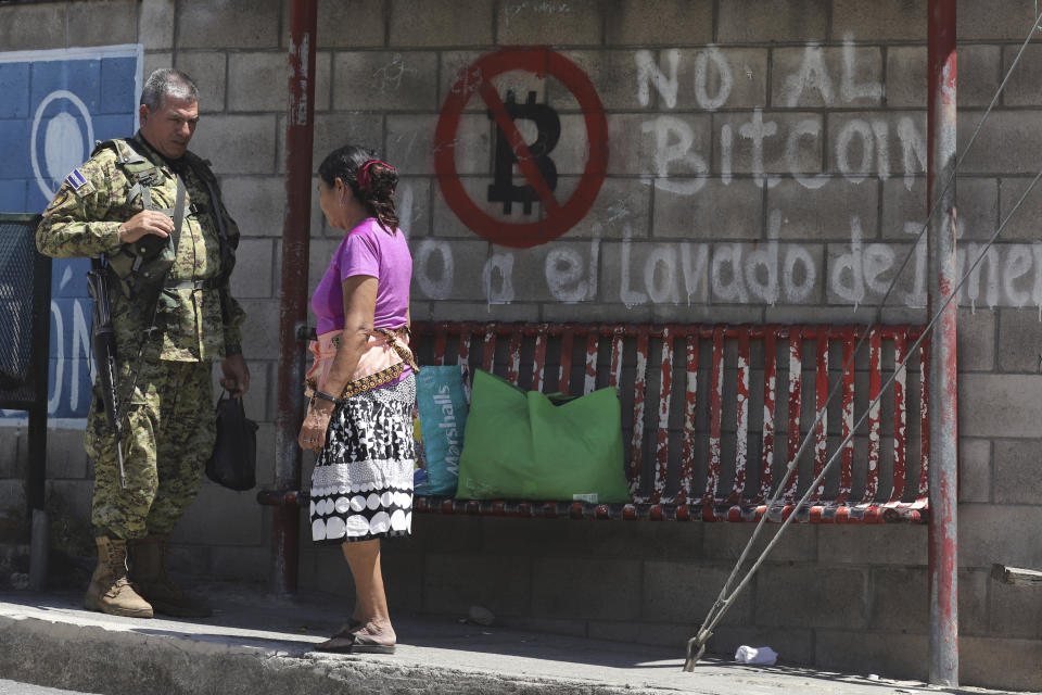 Un soldado habla con una vecina en una parada de autobús cubierta con graffiti en contra del uso del Bitcoin, antes de unas elecciones municipales en San José Las Flores, El Salvador, el miércoles 28 de febrero de 2024. El Salvador celebró sus elecciones municipales el 3 de marzo. (AP Foto/Salvador Meléndez)