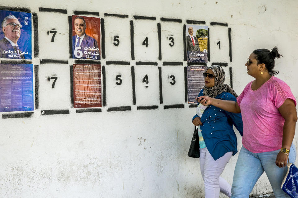Women walk past a wall of campaign posters in Tunis, Tunisia, Monday, Sept. 2, 2019.Tunisia's 26 presidential candidates have launched their campaigns in a political climate marked by uncertainty, money laundering allegations and worries about violent extremism. (AP Photo/Hassene Dridi)