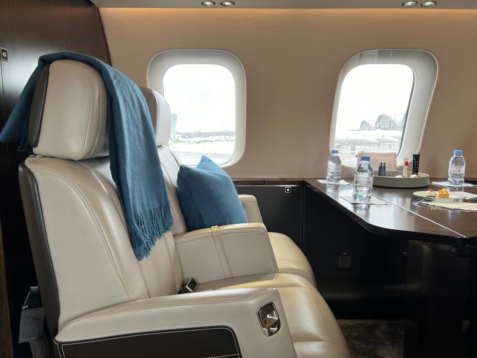 Two large cream leather seats with a blue blanket and cushion next to a table with water bottles, on board a VistaJet Bombardier Global 7500