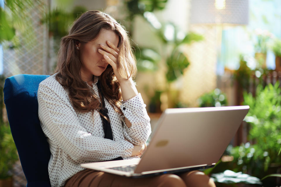 stressed trendy middle aged housewife with long wavy hair in the modern living room in sunny day using laptop while sitting in a blue armchair.