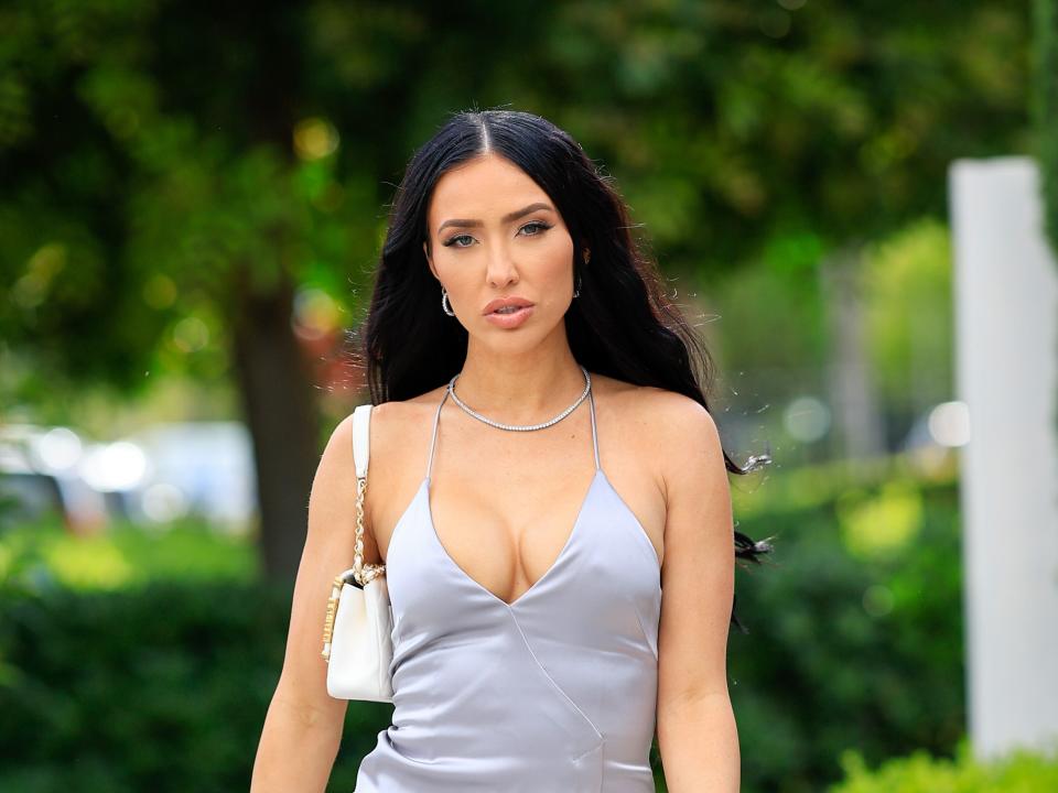 Bre Tiesi walking along a suburban sidewalk while wearing a form-fitting silver dress with a longer skirt on one side, thin straps, and a low cut.