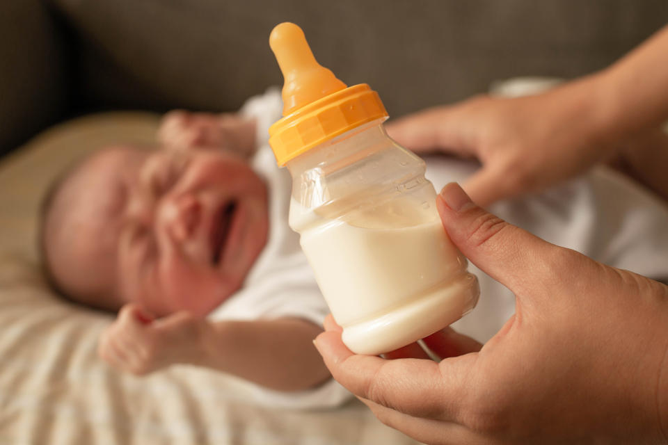 An infant cries as a mother holds a full bottle of formula by its side.