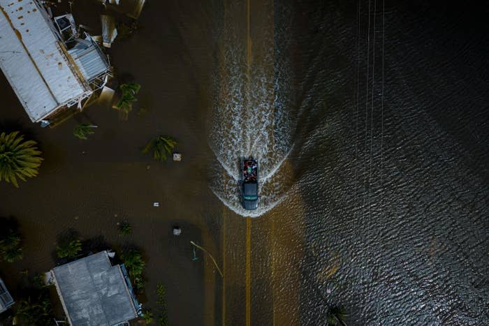 An aerial view of a truck driving down a flooded area, causing waves as it progresses with mobile homes, sunlight reflects off the ripples in the water