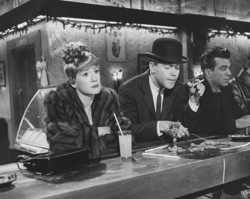 Shirley MacLaine in a fur coat, Jack Lemmon in a bowler hat, and another man sit at a bar counter in a scene from an old movie