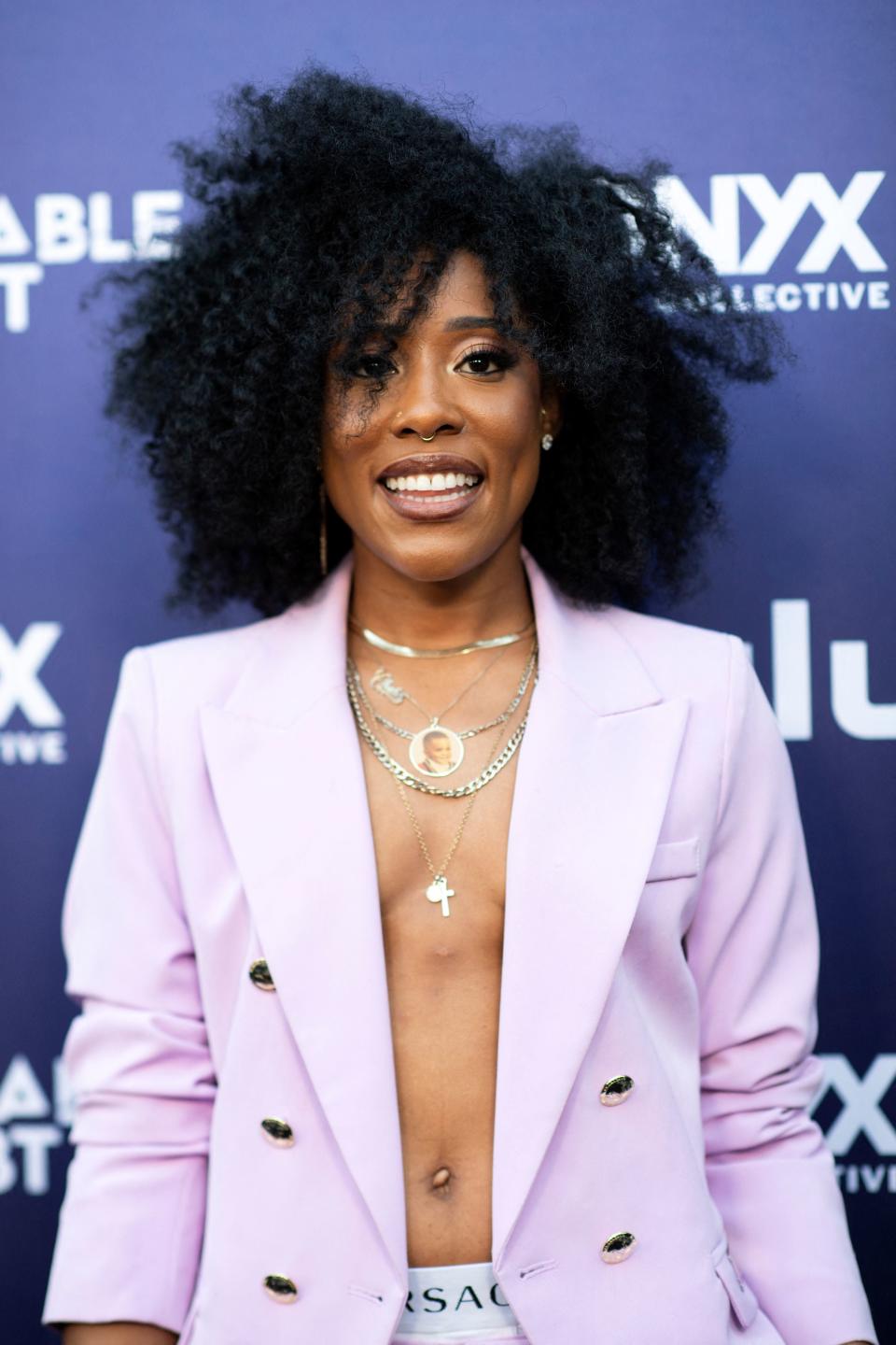 Jonica wearing a lavender blazer with no shirt underneath and layered necklaces, smiling at an event