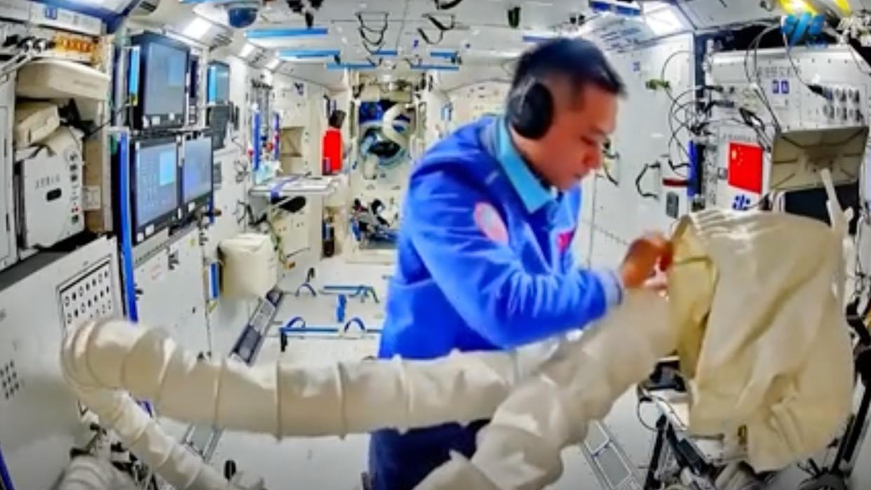  An astronaut in a blue flight suit examines cargo in a white bag aboard a white-walled space station. 