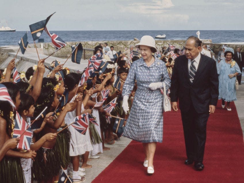 Queen Elizabeth II, wearing a blue-and-white outfit with a white safari helmet-style hat, escorted by an unspecified dignitary alongside a crowd of children waving British flags in Honiara, Solomon Islands, 18th October 1982. Queen Elizabeth II is on a Commonwealth Tour of South Pacific nations.