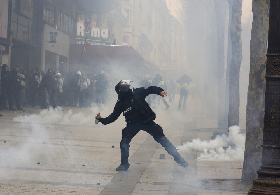 A riot police officer throws a tera gas canister onto protesters during a yellow vests demonstration on the Champs Elysees avenue Saturday, March 16, 2019 in Paris. French yellow vest protesters clashed Saturday with riot police near the Arc de Triomphe as they kicked off their 18th straight weekend of demonstrations against President Emmanuel Macron. (AP Photo/Christophe Ena)