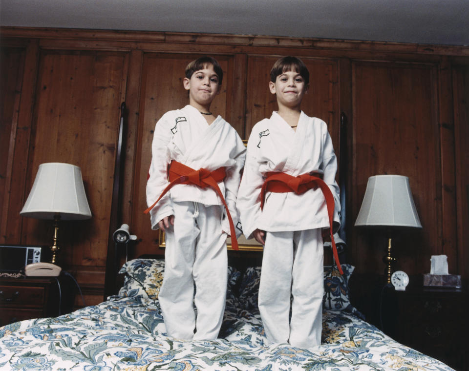 Twin boys, each wearing a white karate gi with red belts, stand on a bed in a room with wooden panel walls and lamps on both sides