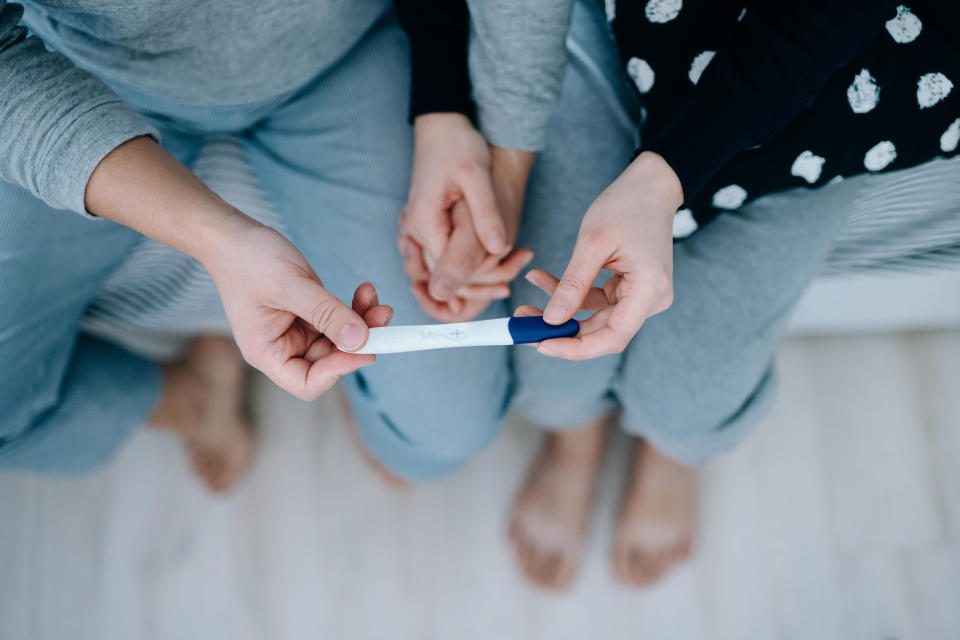 Overhead view of an affectionate young Asian couple sitting on the bed, holding hands and holding a positive pregnancy test together. It's finally happening. The long-awaited news. Life events, fertility and family concept