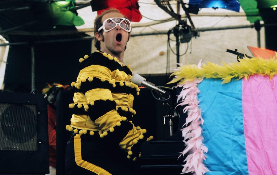 elton john sings while sitting and playing a piano, he wears a bee costume with glasses
