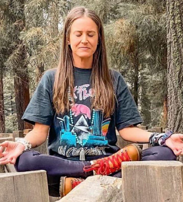 Holly Courtier meditates in the woods while wearing hiking boots and attire. 