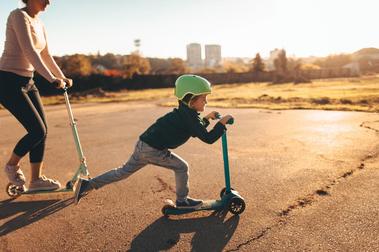 Photo of a cheerful little boy riding a push scooter, with his mom right behind him