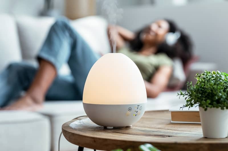 Shot of essential oil aroma diffuser humidifier diffusing water articles in the air while woman listening music lying on coach.