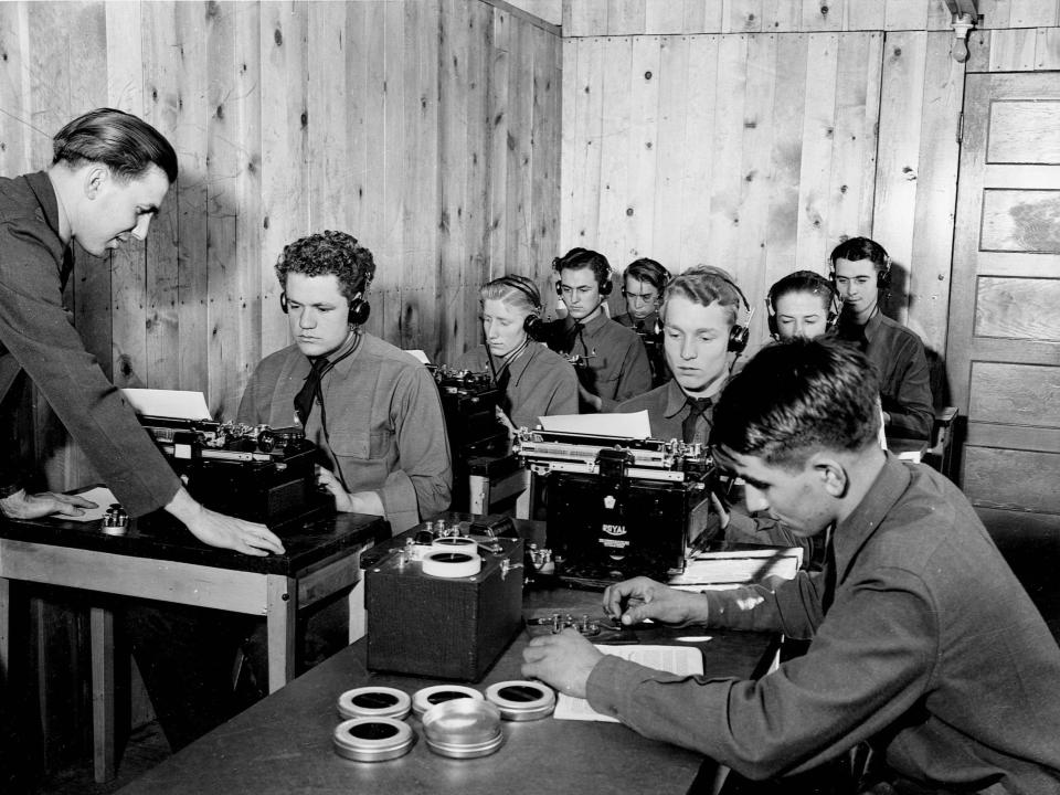 A black and white photo of young men at type writers with headphones on.