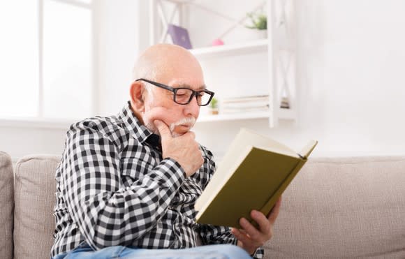 Senior man reading a book on a sofa, with a hand against his chin.