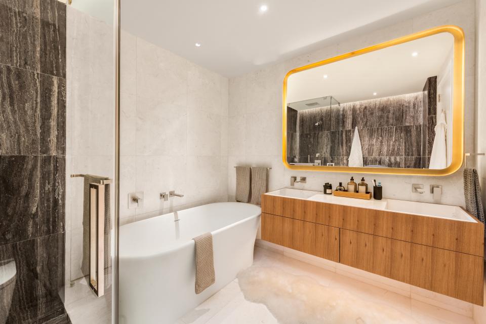 The master bathroom features an oak vanity and a brown travertine shower (at left). Cream-colored marble wraps the walls and floor.