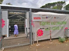 A woman wearing a purple hospital gown can be seen through a metal fence. A large colorful sign reads 'Centro Vaccinazioni Anti-Covid 19' in green letters