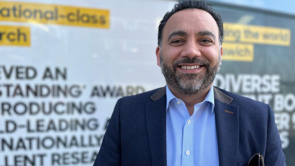 A man in a blue jacket and shirt smiles, behind him are inspirational phrases in the windows of a university building