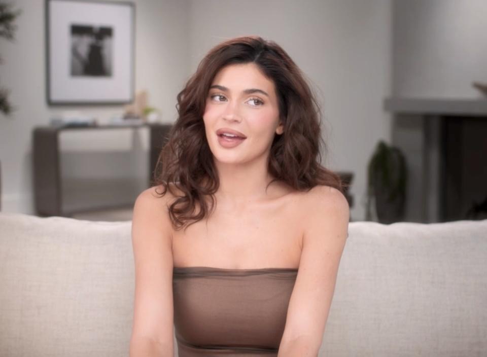Kylie Jenner sits on a couch wearing a strapless top and speaking during a confessional