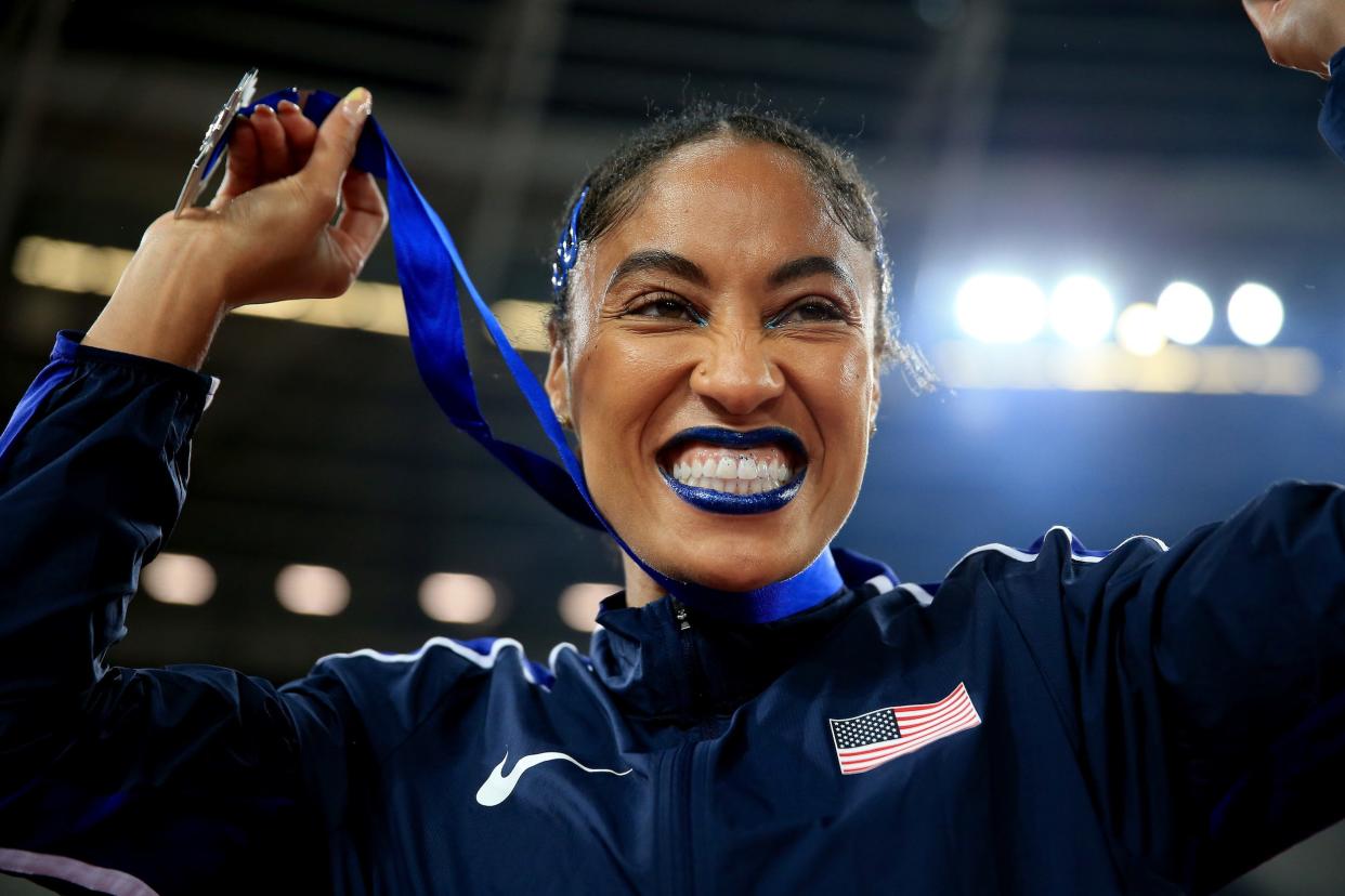 Queen Harrison-Claye in blue makeup, a blue Team USA jacket, and blue hairclips, holds up a blue medal