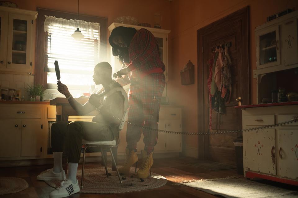 Two characters in a kitchen, with one character seated and the other holding a mirror and grooming their hair. Sunlight streams in from the window