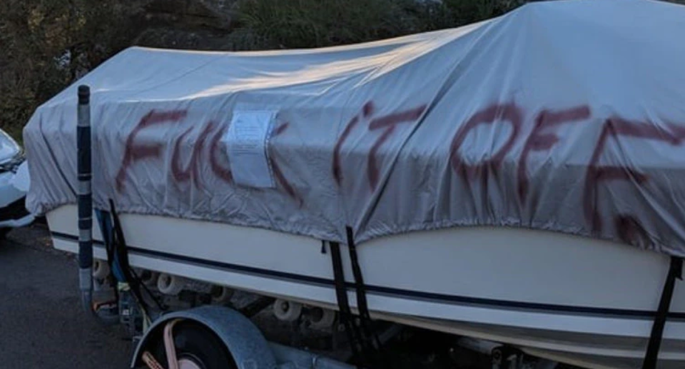 An angry message spay painted in red paint is seen on the tarp covering of a boat parked on the side of the road.