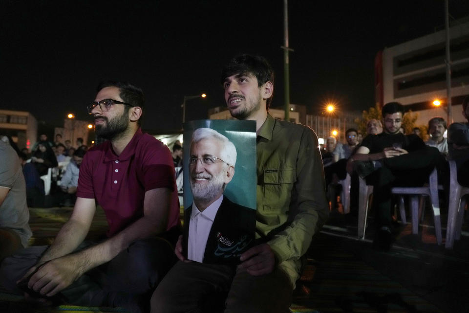 People watch a debate of candidates of June 28, presidential election on a giant screen as a supporter of candidate, Saeed Jalili, holds his poster in downtown Tehran, Iran, Monday, June 17, 2024. The election comes at a time of heightened tensions between Iran and the West over its arming of Russia in that country's war on Ukraine. (AP Photo/Vahid Salemi)