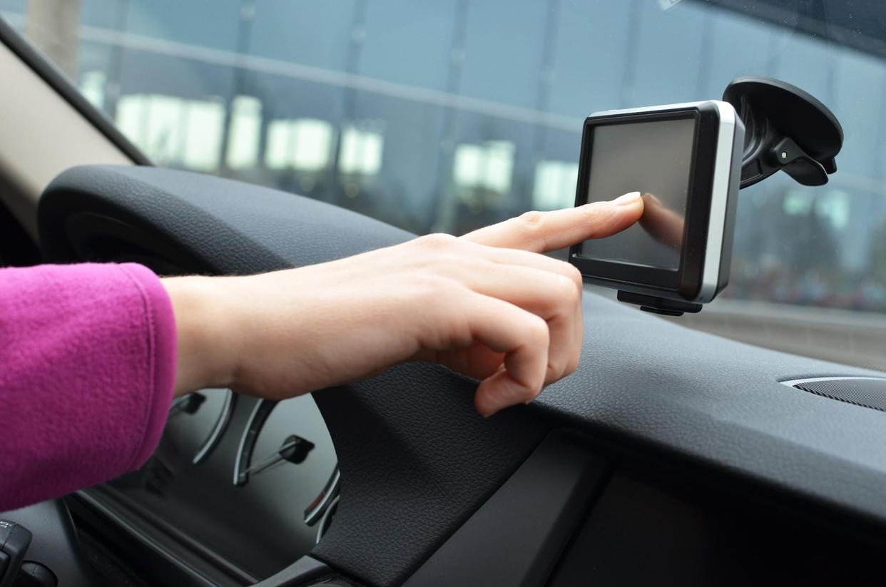 Female driver's hand touching GPS device hanging on windshield in car, dark grey dashboard, woman's arm is covered in a violet fleece, with a blurred background of a building