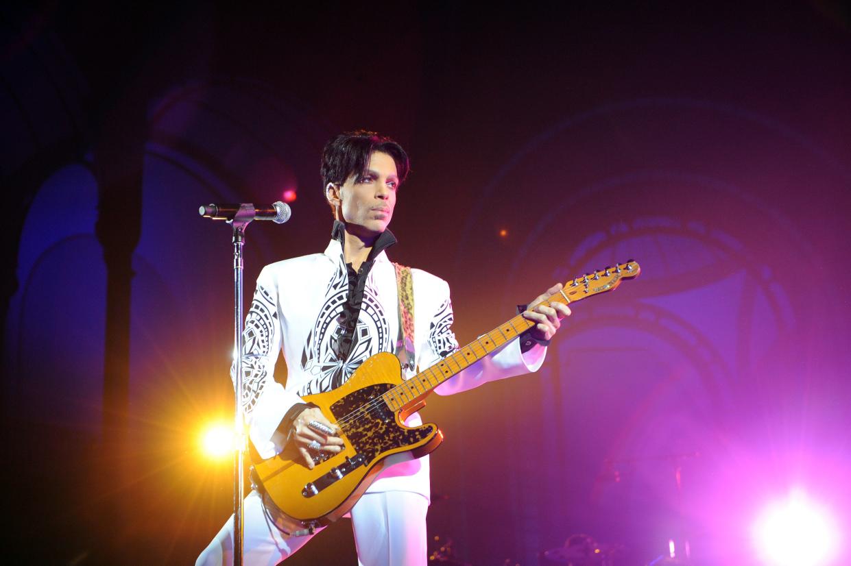 Prince on stage with his guitar and he looks dazzling in white and black print shirt and pant to match. 