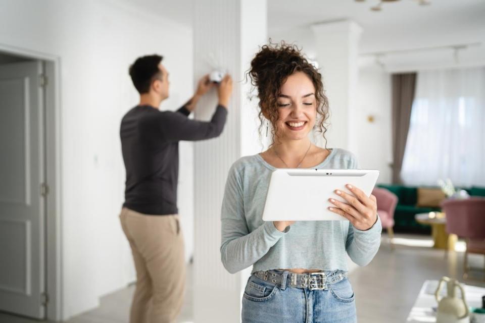 A woman smiles while looking at a tablet's screen; a man behind her presses buttons on a wall-mounted home security system.