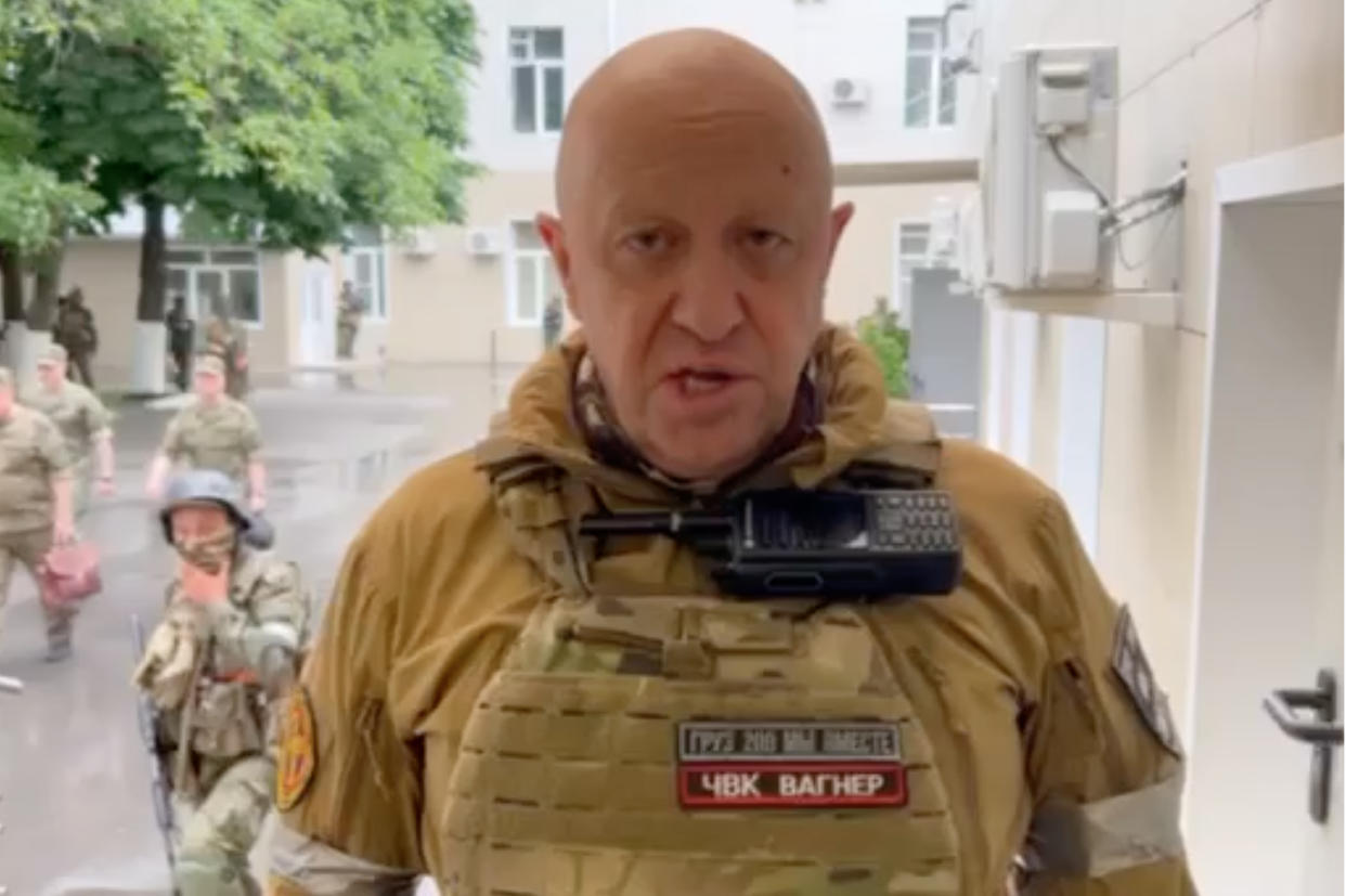 Prigozhin stands on a street with soldiers behind him as he looks at the camera. 