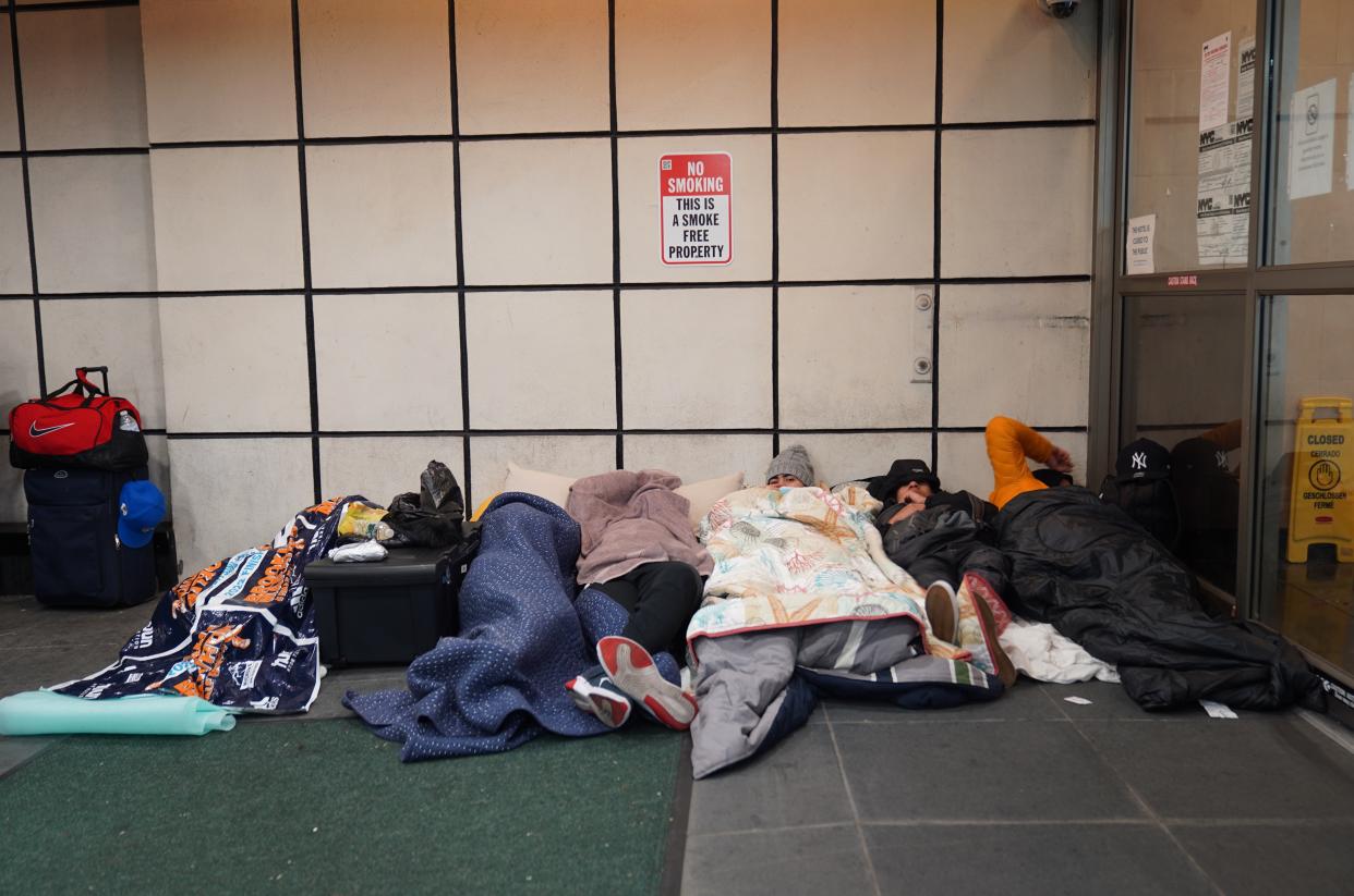 Seven people sleep on the ground, with luggage nearby, under a sign that says: No Smoking, This Is a Smoke-Free Property..