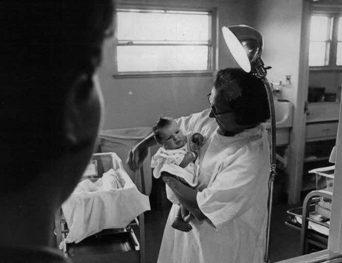 A nurse cares for a newborn in a hospital nursery while a woman watches through a window