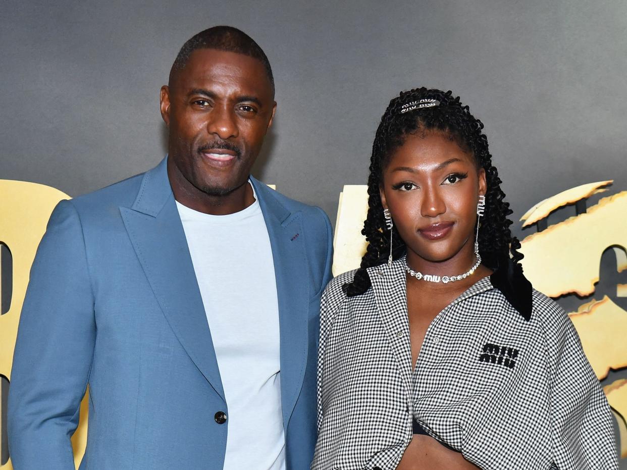 idris elba and his daughter isan on the red carpet for beast posing together. elba is smiling and wearing a light blue suit with a white t-shirt under an unbuttoned jacket. isan is smiling with her mouth closed, wearing a  two-part grey suit that's only buttoned in the middle, exposing her midriff