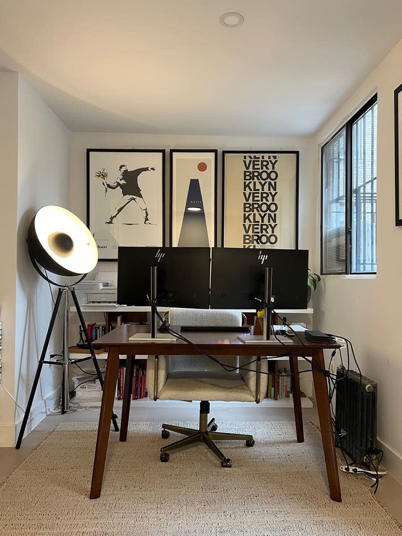 Wood desk with two monitors next to large tripod floor lamp in beige office with hung artwork.