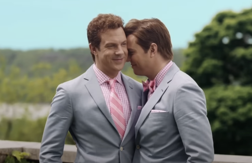 Two men in light suits with pink checkered shirts and pink ties stand close outdoors, smiling and touching foreheads