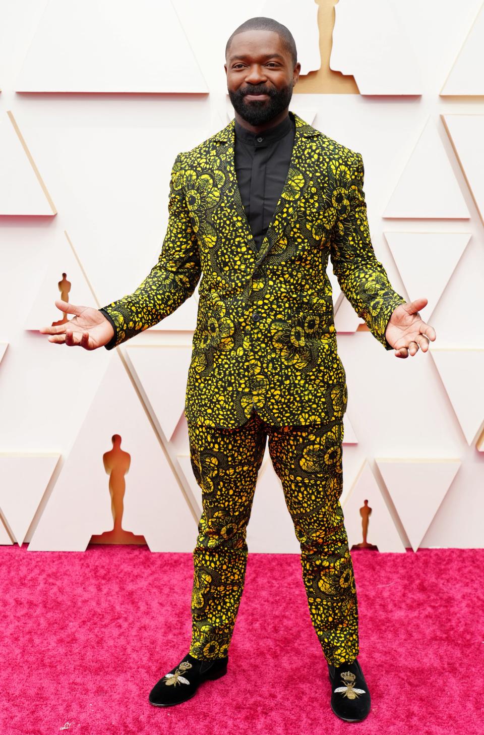 David in a green-black suit with a neon yellow floral print on the jacket and pants. He has a black button down on and black velvet loafers with embroidered bees on them.