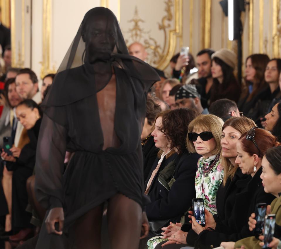 Anna Wintour and other attendees watching a runway show; model in sheer black dress and veil walks towards the camera