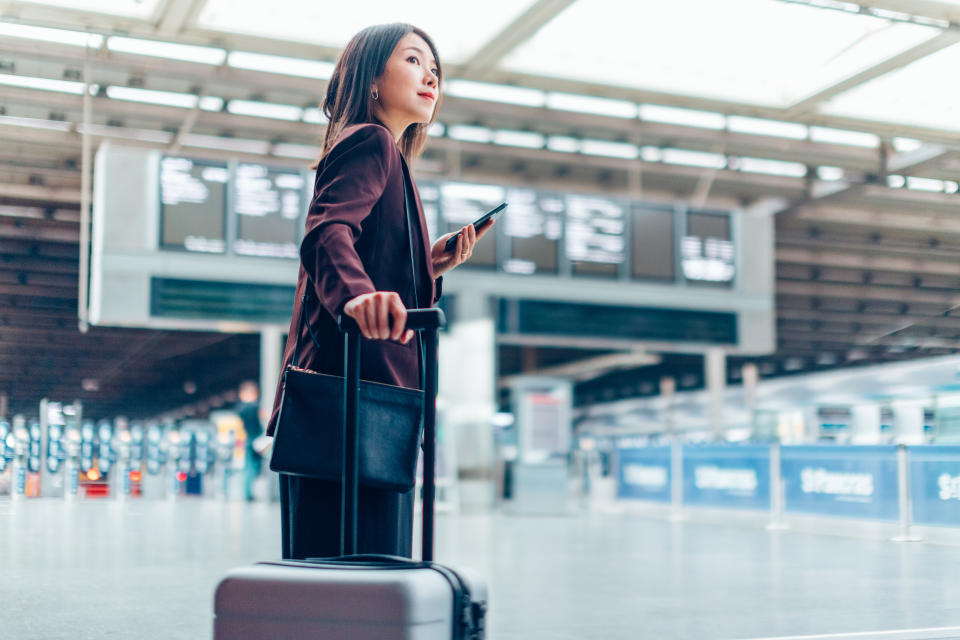 Young Asian businesswoman holding phone and carrying suitcase in airport departure area