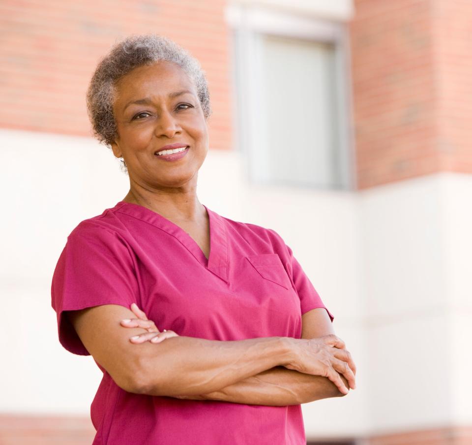 A woman wearing scrubs stands outside a building, smiling with arms crossed