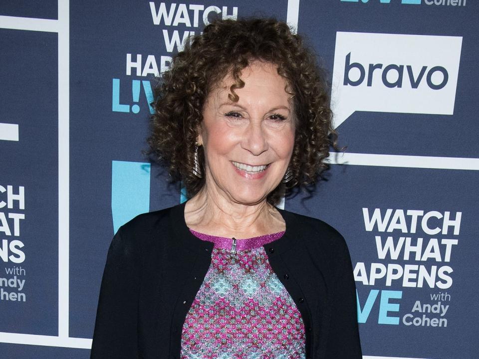 Rhea Perlman wearing a multicolored dress with yellow, red, purple, and blue with a black cardigan in front of a blue background