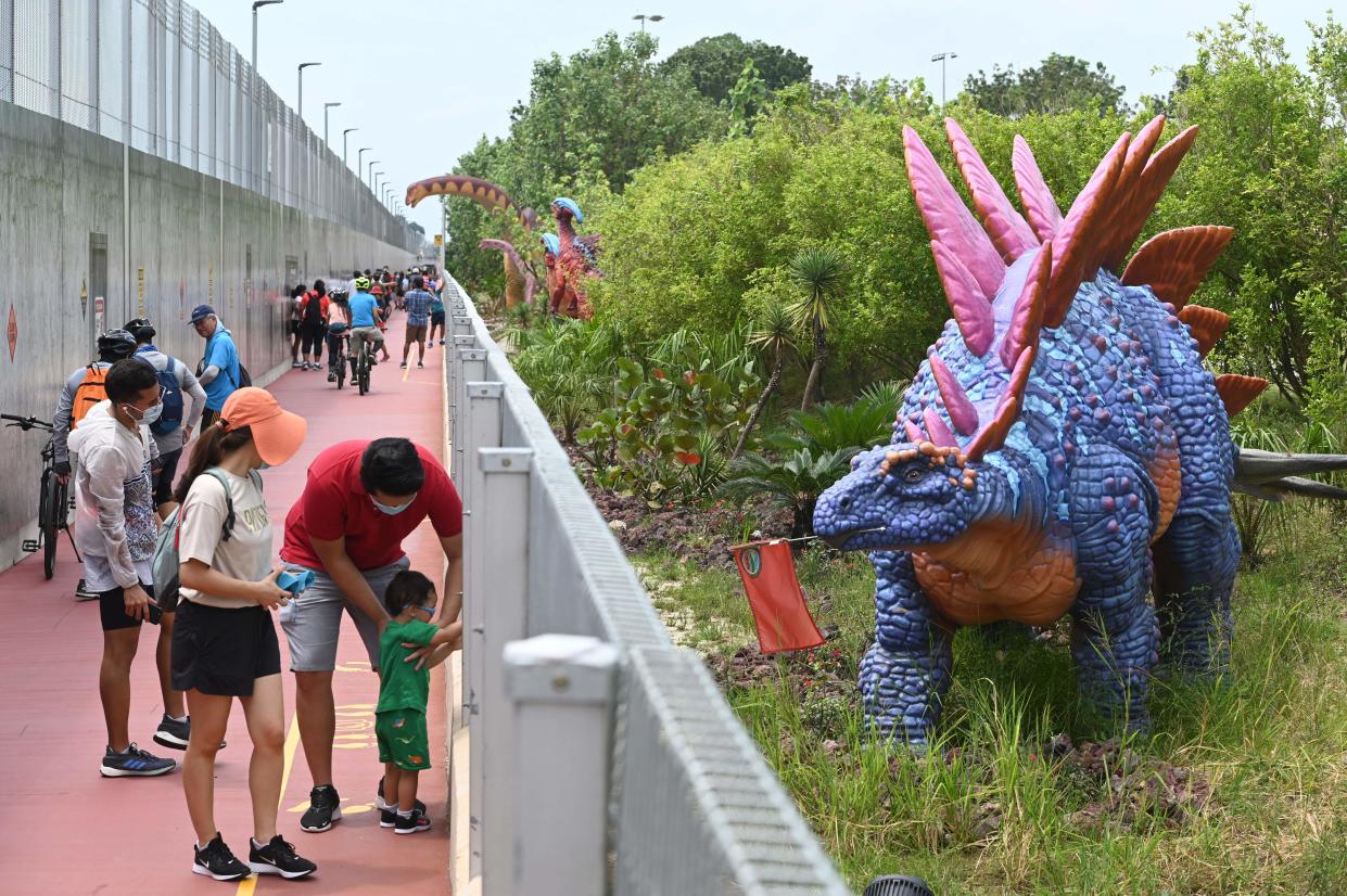 People stop to take photograph of a life-size dinosaur model displayed along "Changi Jurassic Mile" a leisure path way in Singapore on October 13, 2020. - The new leisure attraction with dinosaur displays connecting Changi Airport terminal 4 and East Coast park allows people to cycle or jog to the airport. (Photo by Roslan RAHMAN / AFP) (Photo by ROSLAN RAHMAN/AFP via Getty Images)